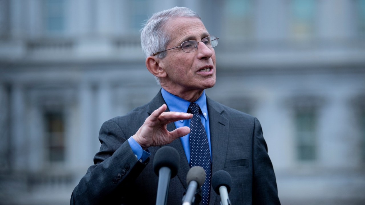 dr anthony fauci stands in front of mics at a podium