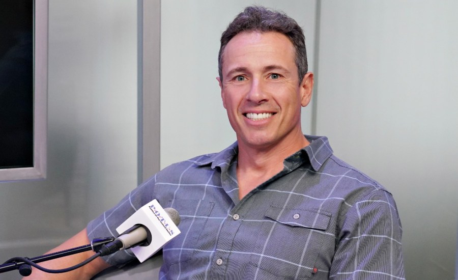 chris cuomo sits in front of a mic, smiling, with a short sleeve button down shirt