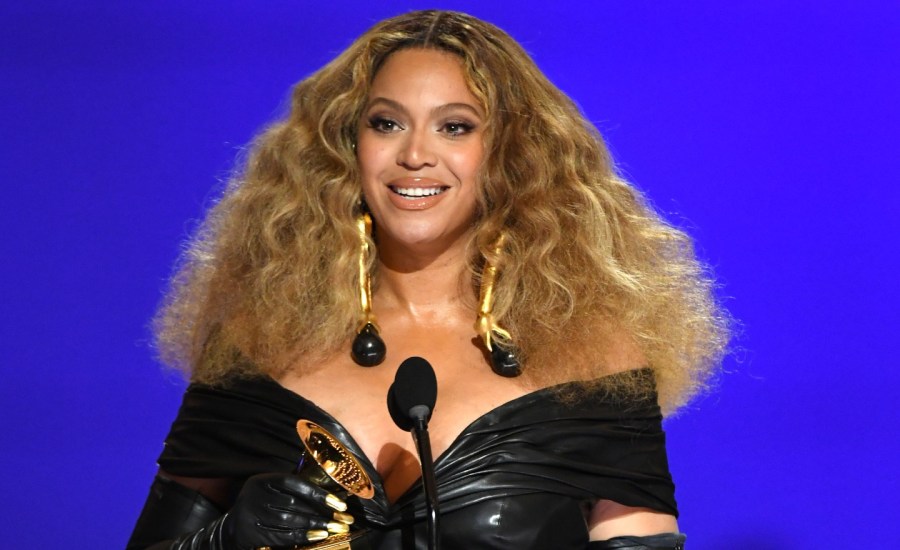 Beyonce Knowles-Carter smiles, holding a Grammy award, on-stage in front of a mic, in a black off-shoulder dress, at the Grammy Awards Ceremony.