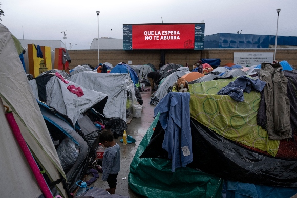 A migrant child stands amid tents at an improvised camp near a screen reading May the wait don't bore you outside El Chaparral crossing port