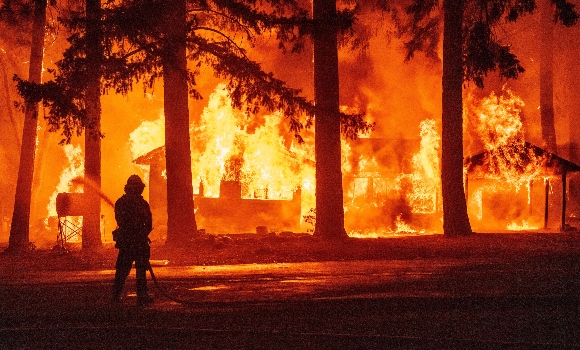 A firefighter sprays water on a propane tank as a home burns due to the Dixie fire in the Indian Falls neighborhood of unincorporated Plumas County, California