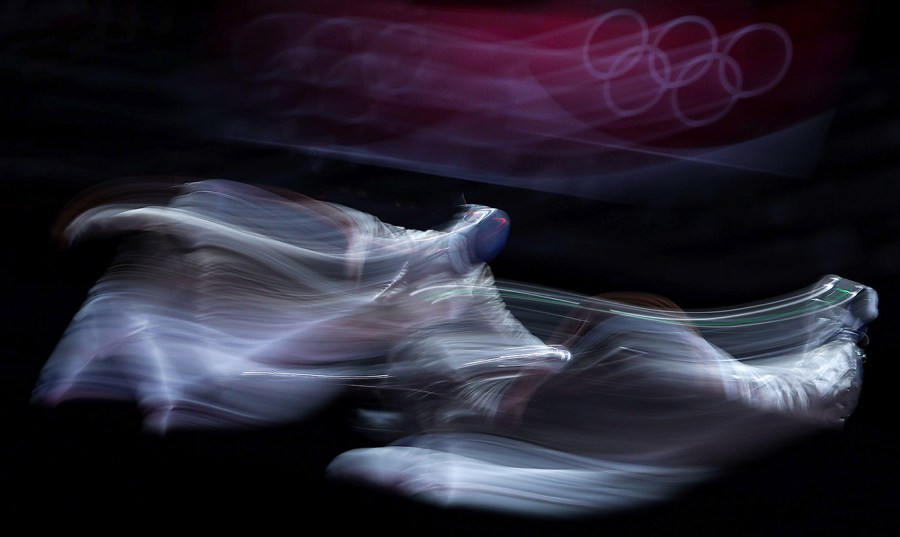 The blurred forms of sabre semi-finals team competitors are seen in front of an also blurred Tokyo Olympics logo