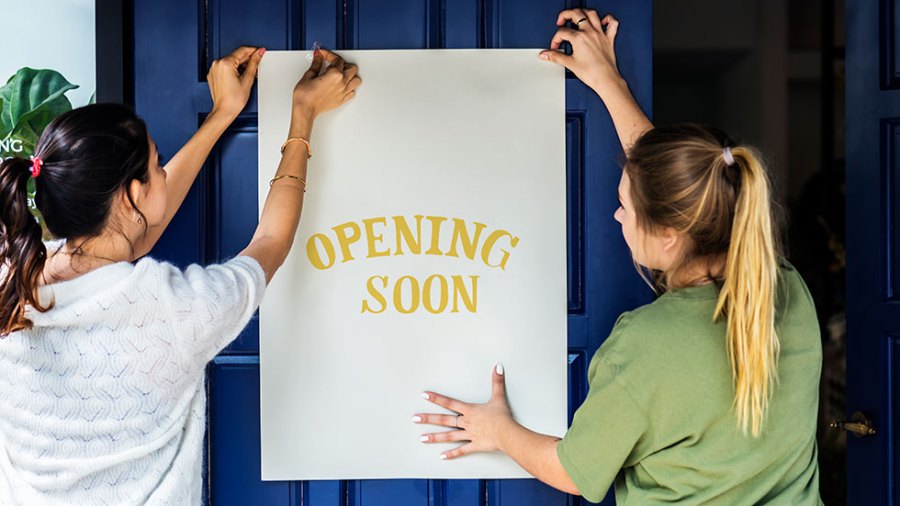 Two women hang a sign that says "Opening Soon"