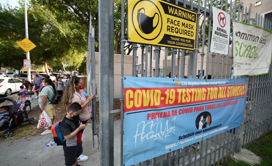 some adults and students waiting at gate outside of school, gate has signs for wearing a mask and testing for COVID-19