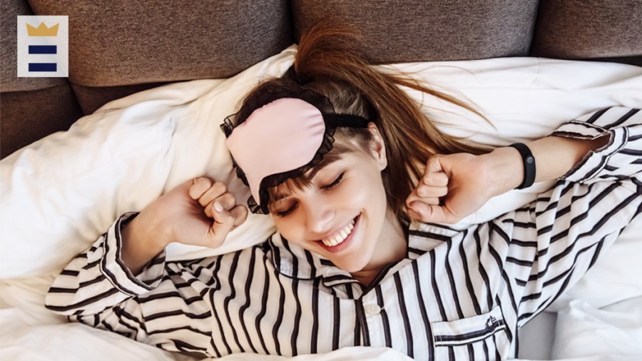 Smiling person wearing striped pajamas and a pink eye mask stretches in their bed.