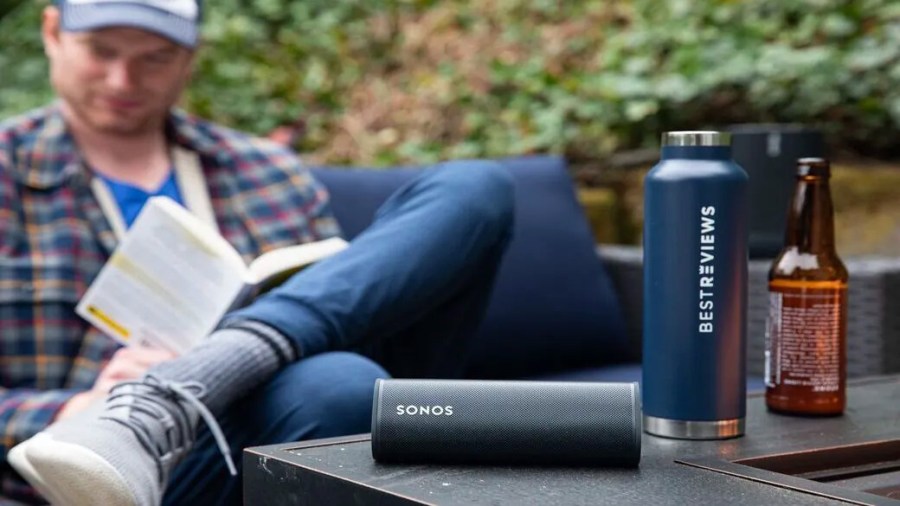 Man reading a book beside a table holding a portable speaker and reusable water bottle