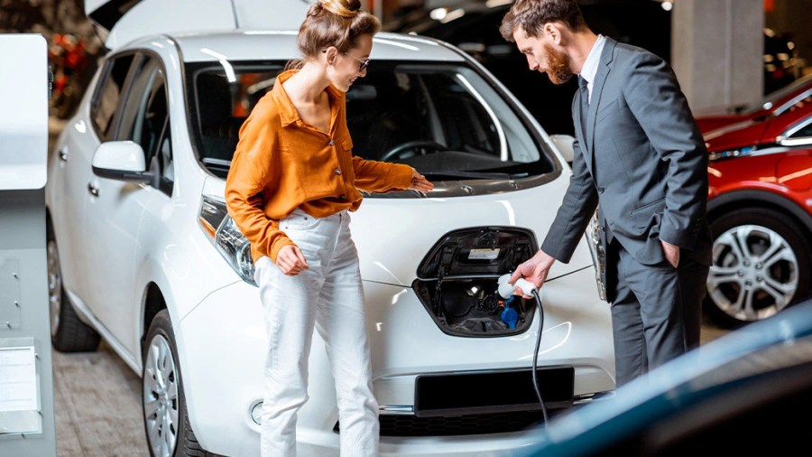 Man plugs an electric car charger into his car's charging port while a female friend watches