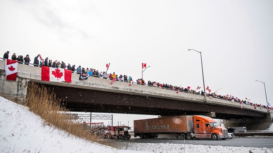 Protesters stand on a bridge in Canada as trucks drive under.