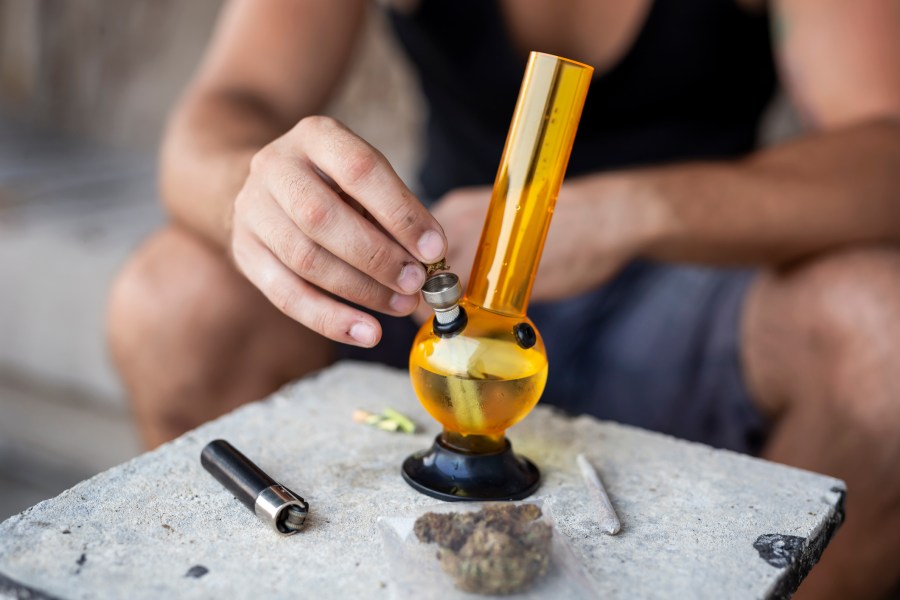 Young man smoking pot using bong; detail of male hand filling up bong with cannabis, getting it ready for use