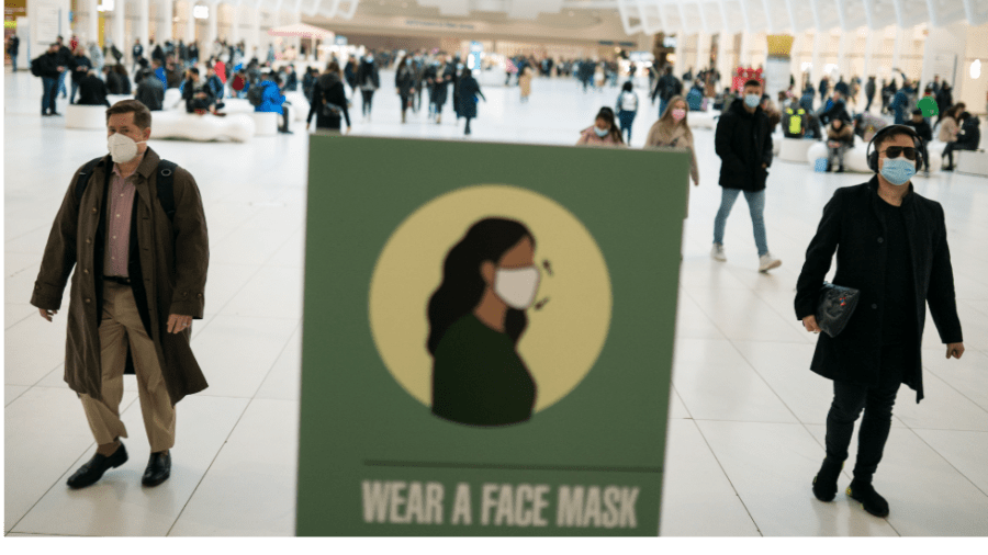 Two masked travelers walk beside a green sign featuring a masked woman that reads "Wear a face mask"