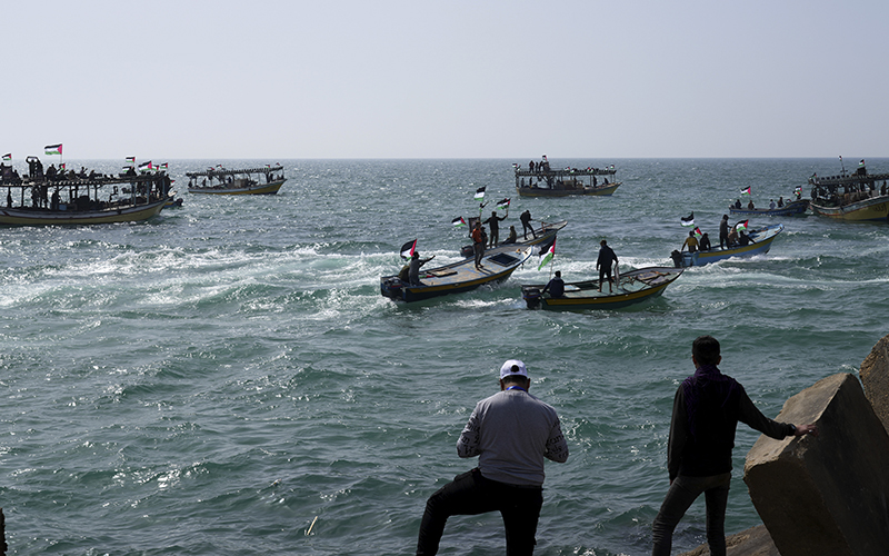 Palestinian watch fishermen while ride their boats and wave their national flags over blue ocean waters