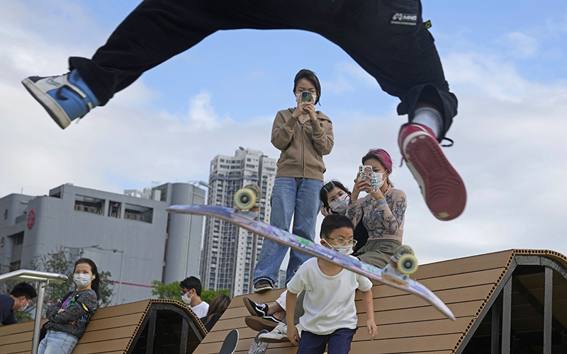 A skateboard trick is seen from below and behind as a crowd of people wearing masks look on