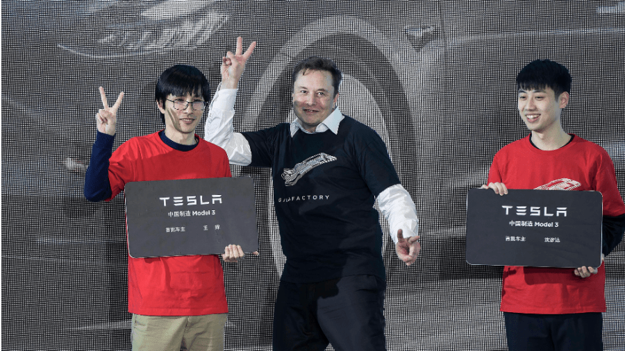 Elon Musk, wearing a black t shirt over a white collared shirt, poses between two Tesla owners in Tesla's Shanghai factory.