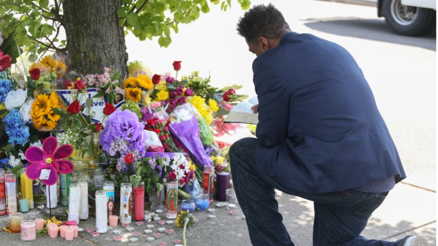 A man wearing a blue suit and white sneakers kneels at a makeshift memorial, reading the Bible.