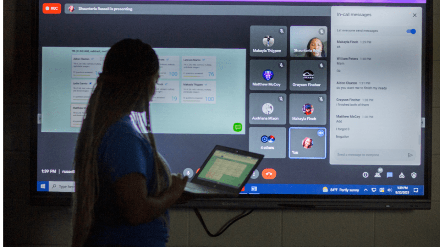 A woman holding a laptop stands in front of a large screen displaying a video conference.