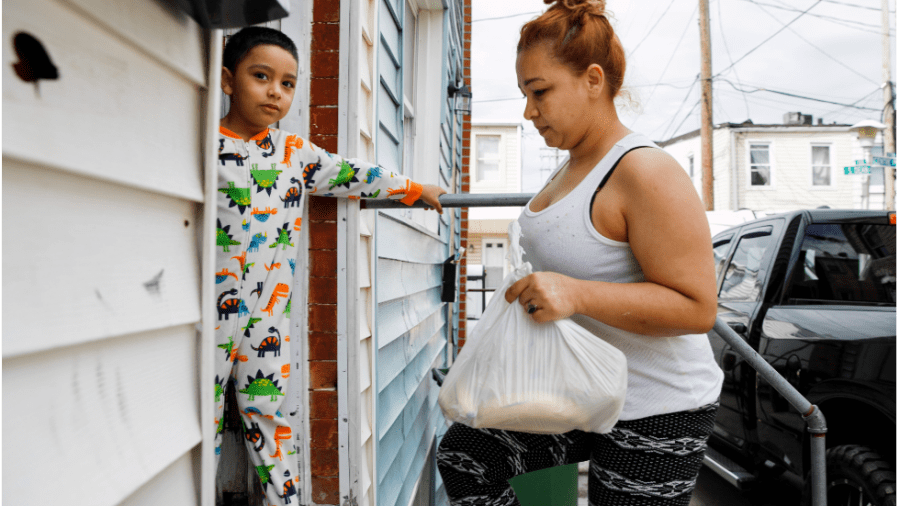 A latino woman in a white top carrying a white bag walks up the steps to her home. Her son, wearing colorful dinosaur pajamas, stares into the camera.