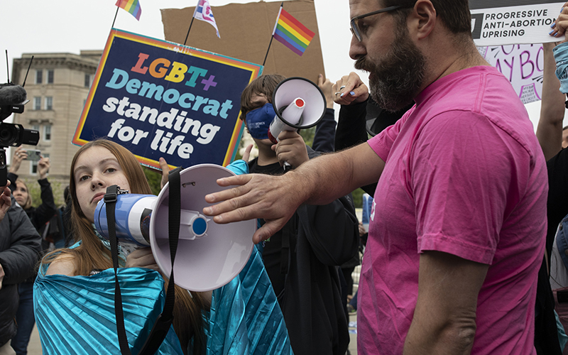 A person puts up their hand over a counterprotestor's microphone with a sign that reads "LGBT+ Democrat standing for life"