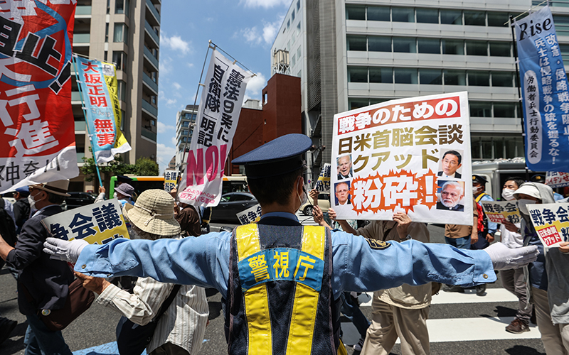 People hold signs and chant slogans during a protest against the Quad summit as a police officer keeps protestors at bay