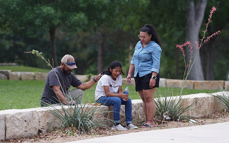 A girl cries, comforted by two adults, outside as she sits on stone blocks