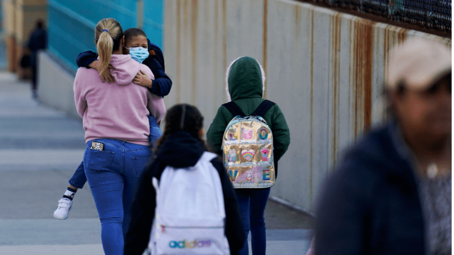 A woman carries a child wearing a mask as two other children wearing backpacks walk behind them.