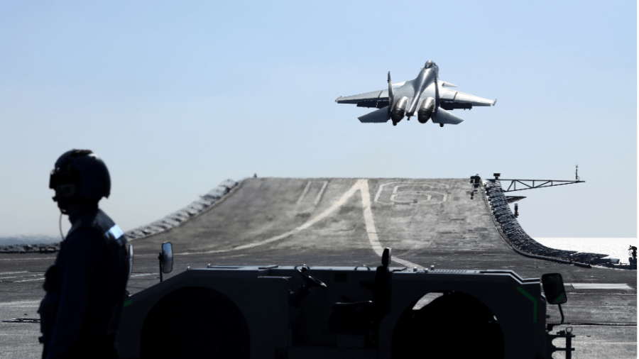 A Chinese fighter jet takes off from an aircraft carrier as a soldier stands nearby.