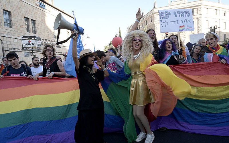 People pose in front of a rainbow flag, backed by a crowd with signs, in Jerusalem's 20th LGBTQ Pride Parade