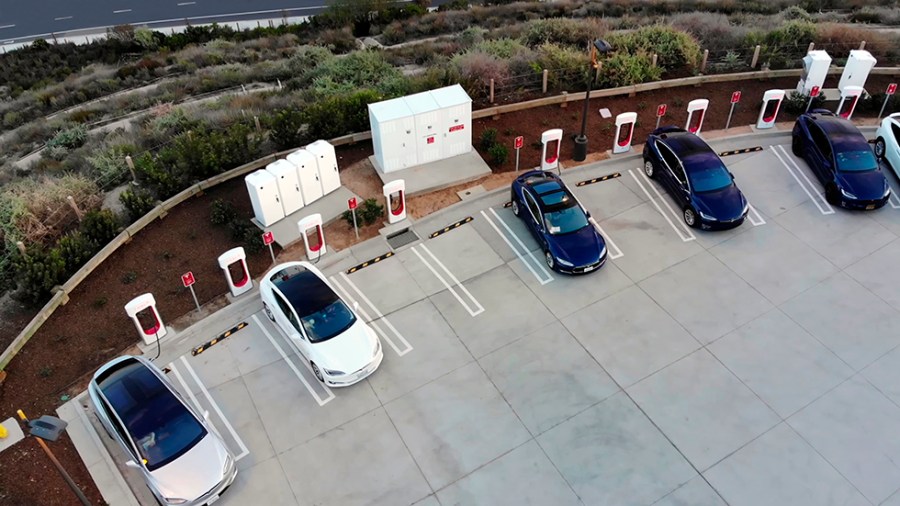 View of a white Tesla Model X, a white Tesla Model S and three blue Tesla Model 3s, parked in a parking lot and charging at a Tesla Supercharger electric charging station.