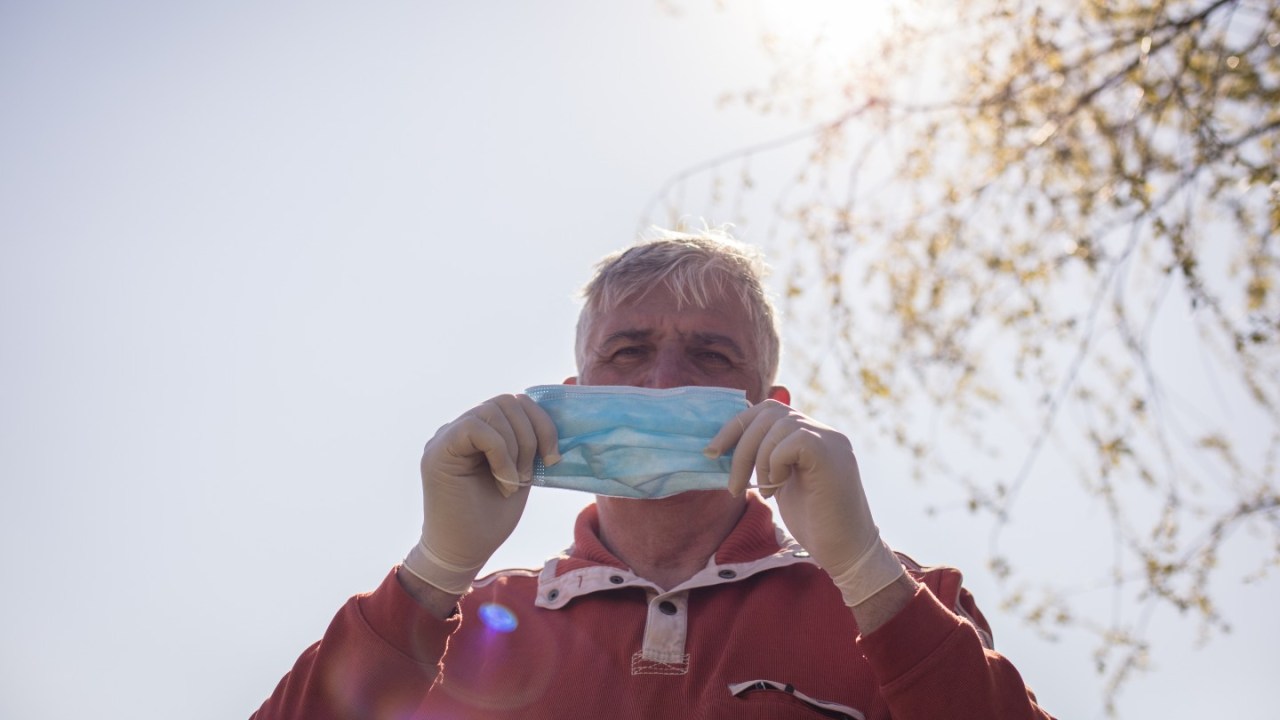 an older adult man standing outside putting on a face mask with sun and tree behind