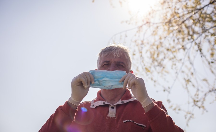 an older adult man standing outside putting on a face mask with sun and tree behind