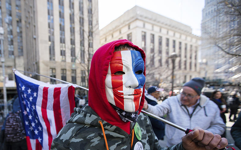 A man wearing a patriotic colored Guy Fawkes mask and holding an American flag, joins a small group of protesters near the Manhattan District Attorney's office