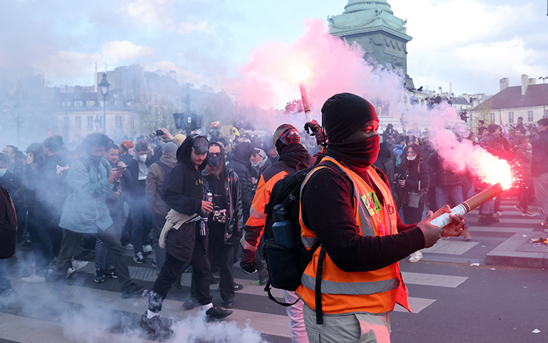 A demonstrator in an orange safety vest holds a lit flare as smoke billows out over the crowd behind them