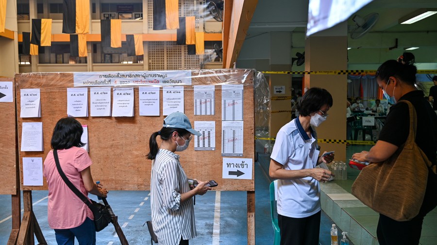 Three people wait in line in front of a poll worker to cast ballots in the Thai general election. There is an information board on wood behind them.