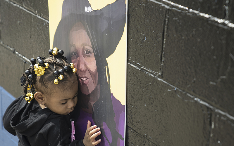 A young girl hugs a purple and yellow portrait of her godmother on a painted cinderblock wall