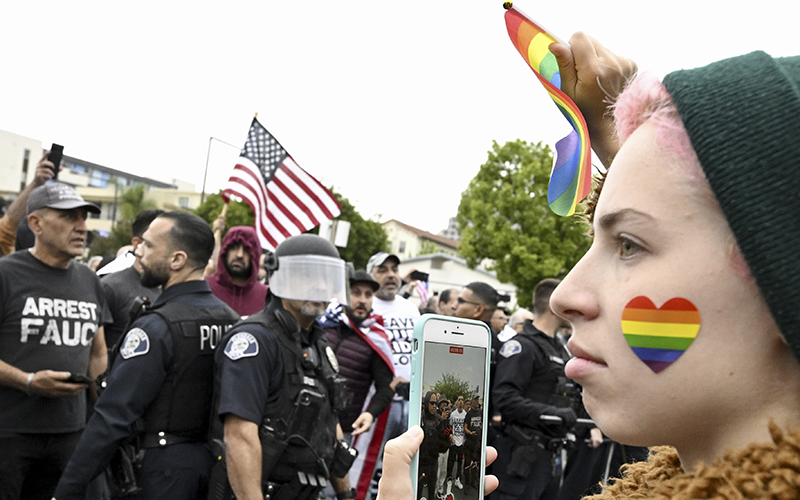 A closeup of a participant with a heart-shaped rainbow on their cheek is shown in the right foreground before a line of police officers who are standing between LGBTQ rights supporters and conservative groups