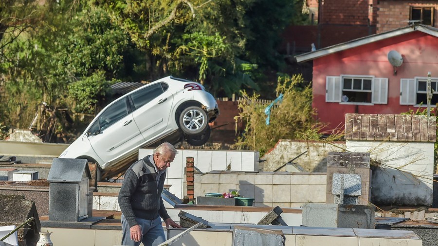 A damaged vehicle lies at an angle over a marble structure in a cemetery while a man walks through the foreground.