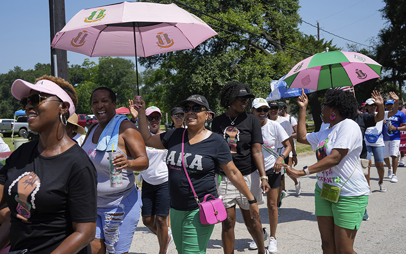 Attendees march in a parade to commemorate Juneteenth, carrying pink and green umbrellas while they laugh and wave