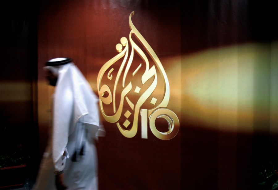 FILE - A Qatari employee of Al-Jazeera Arabic language TV news channel walks past the logo of Al-Jazeera in Doha, Qatar. The pan-Arab news network Al Jazeera has condemned a recent decision by Egyptian authorities to brand some of its journalists as terrorists. The media outlet, which is owned by the Gulf state of Qatar, said that “a number” of its Egyptian journalists and presenters had been added to a list of alleged terrorists published in an official newspaper earlier this month following a ruling by the Cairo Criminal Court. (AP Photo/Kamran Jebreili, File)