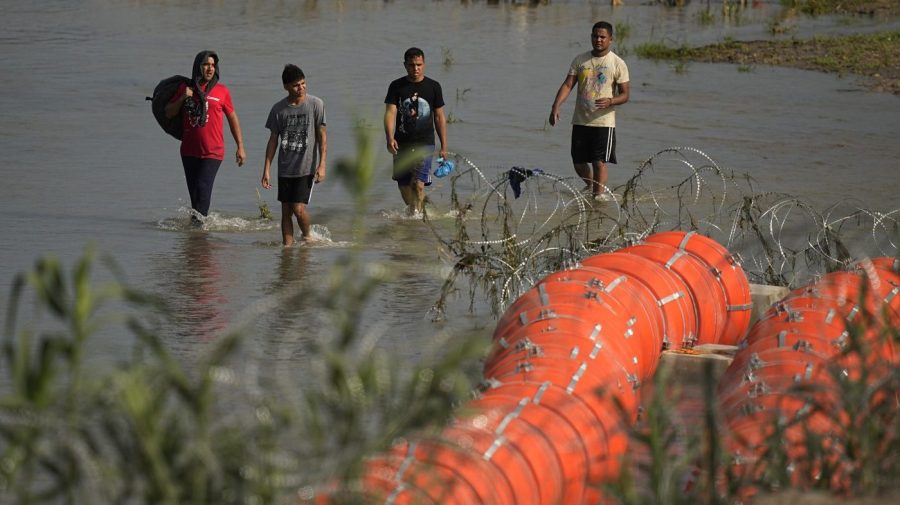 Migrants walk past the site where workers are assembling large buoys to be used as a border barrier along the banks of the Rio Grande in Eagle Pass, Texas, Tuesday, July 11, 2023.