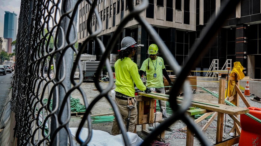 Construction workers in neon yellow attire are seen through a chain link fence close up