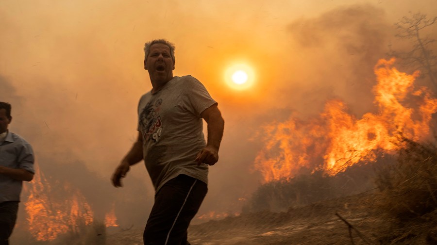 A man stands in the center of the frame while a fire burns brush in the background. Through the smoky haze, the sun can be seen dimly shining.