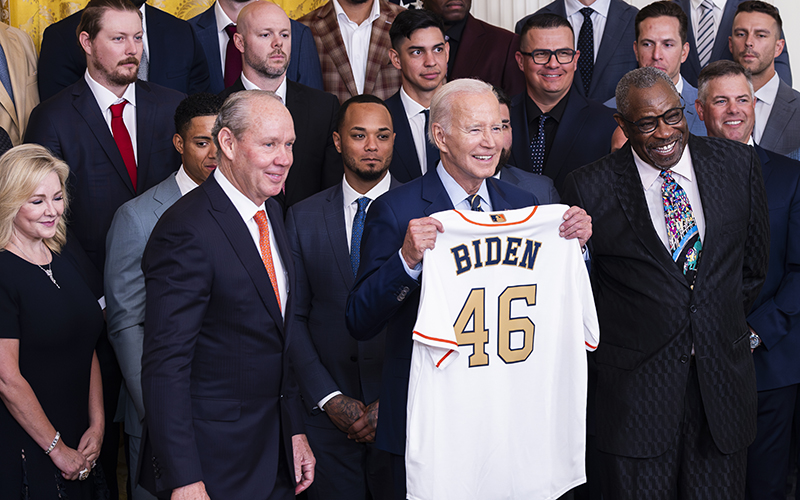 President Biden holds up a Houston Astros jersey that reads "Biden 46." Behind and around him are members of the Houston Astros posing for a photo