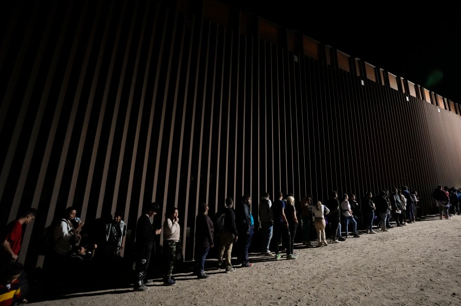 FILE - People line up against a border wall as they wait to apply for asylum after crossing the border from Mexico. Tuesday, July 11, 2023, near Yuma, Ariz. As the number of migrants coming to the U.S.'s southern border is climbing, the Biden administration aims to admit more refugees from Latin America and the Caribbean over the next year. The White House Friday, Sept. 29, released the targets for how many refugees it aims to admit over the next fiscal year starting October 1 and from what regions of the world. (AP Photo/Gregory Bull, File)