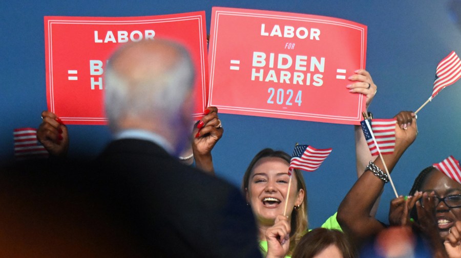 People hold up red signs that read "Labor Biden Harris 2024"; Biden is seen from behind blurred in the background