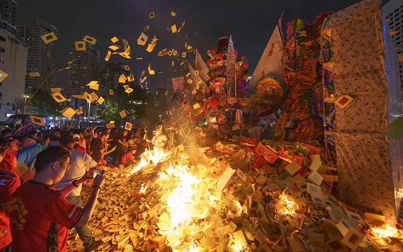 People throw paper offerings into a fire in front of a giant paper statue of a Chinese deity