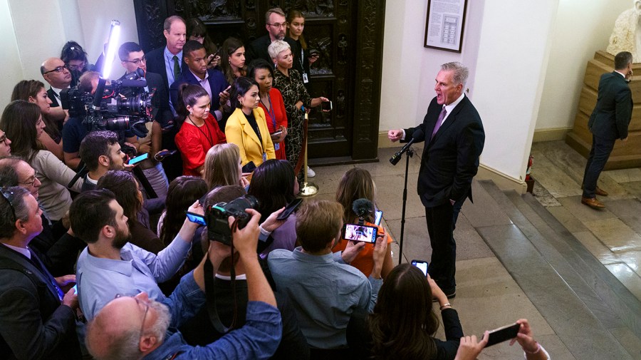 Speaker Kevin McCarthy (R-Calif.) addresses reporters about funding the government. McCarthy is standing in front of a microphone on the right side of the image, and on the left is a group of reporters and photographers standing in a semicircle. The angle of the photo is taken from slightly above the crowd, looking down at the scene.