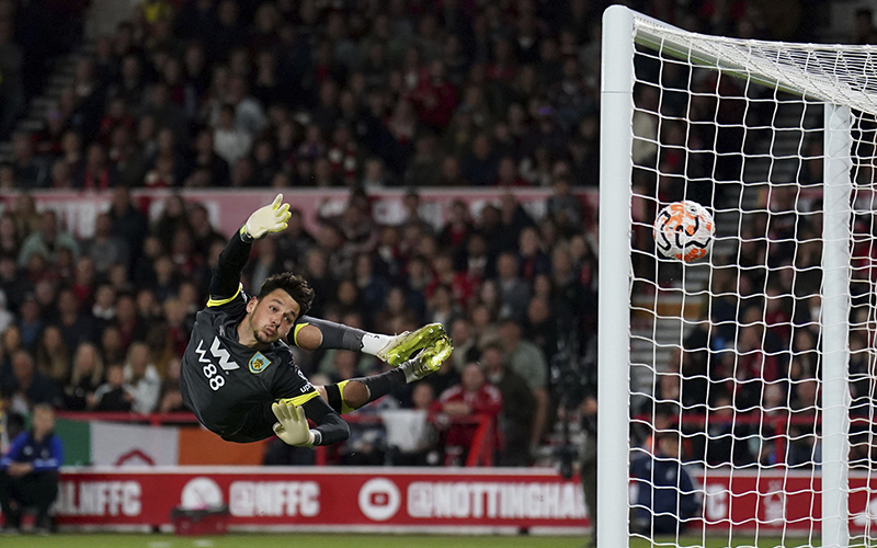 Burnley goalkeeper James Trafford dives in vain as Nottingham Forest's Callum Hudson-Odoi scores during a English Premier League soccer match. On the left side of the image is the goalkeeper, who is in midair. His arms reach toward the ball that has passed him. On the right of the image is the goal net, with the soccer ball in midair, going into the net.