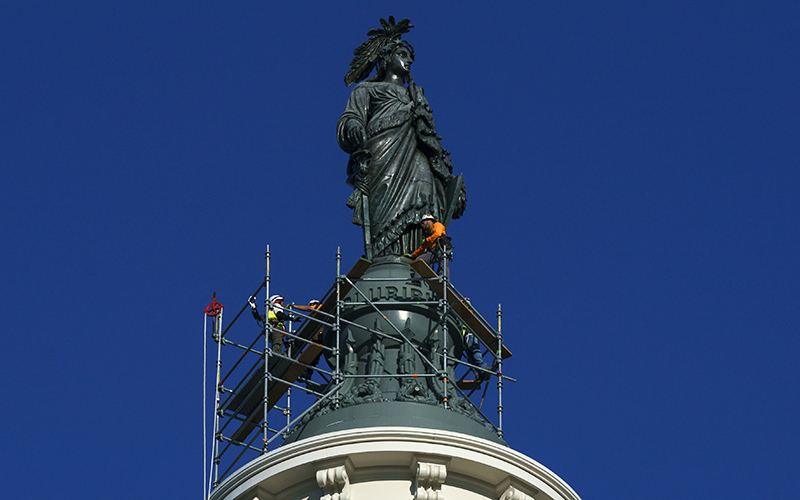 Workers begin a refurbishing project of the State of Freedom atop the Capitol. The statue is in the center of the image, framed against a clear blue sky. Around the base of the statue is a metal scaffolding. Two workers stand on the scaffolding to work on the statue.