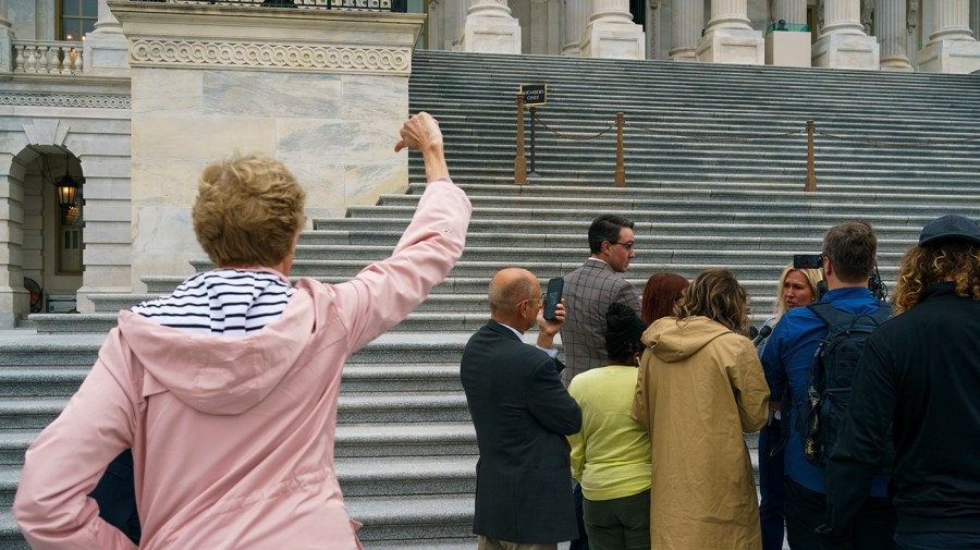 A group of reporters stand in front of the Capitol steps, surrounding Rep. Marjorie Taylor Greene (R-Ga.) to interview her. A little distance away, closer to the foreground, a woman in a pink jacket faces the group with her right hand raised in a thumbs down gesture.