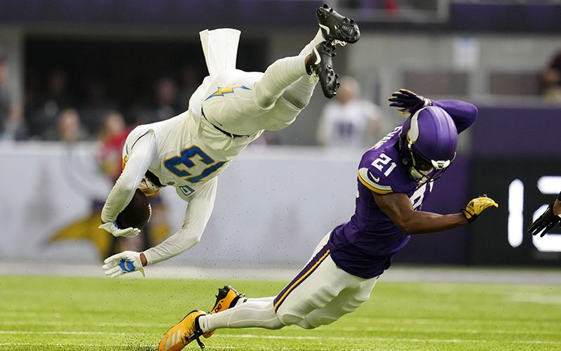 A Los Angeles Charges player is seen in the upper left, suspended in midair as he dives. He has the football tucked in the left hand and the other hand reaches toward the ground. The Vikings player is facing away, about to fall on the field, with his left hand raised behind his helmet