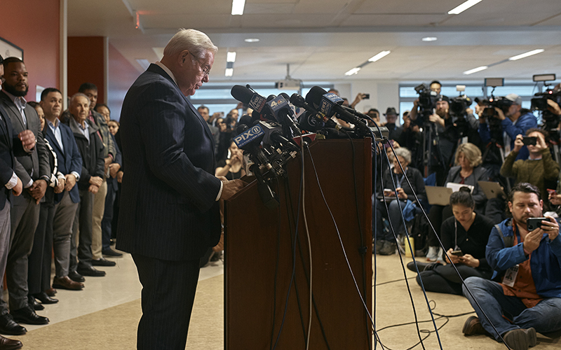 Sen. Bob Menendez (D-N.J.) is seen in profile, standing at a podium that has lots of microphones attached to it. He is looking down to read a statement. In the background and on the right side of the image are members of the media, holding phones,laptops and cameras